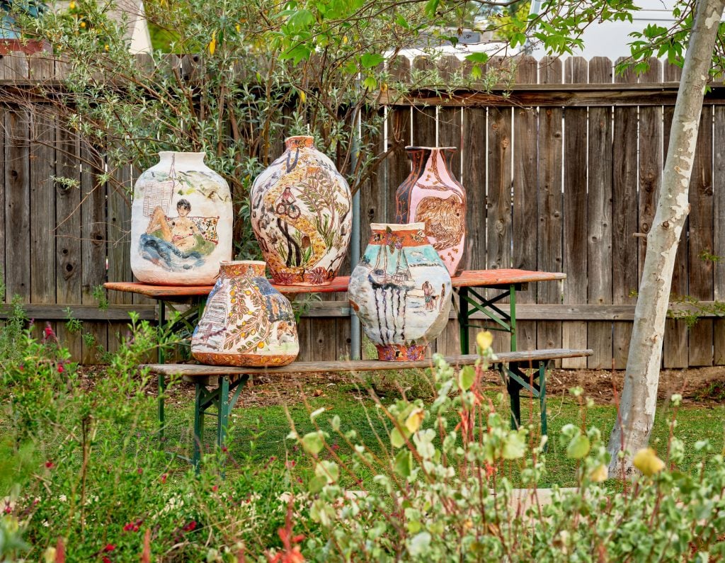 A selection of five colorfully painted and drawn on terracotta vessels by artist Jennifer Rochlin arranged on a table and bench with green metal legs outdoors in front of a wooden fence and surrounded by green grass and other foliage.