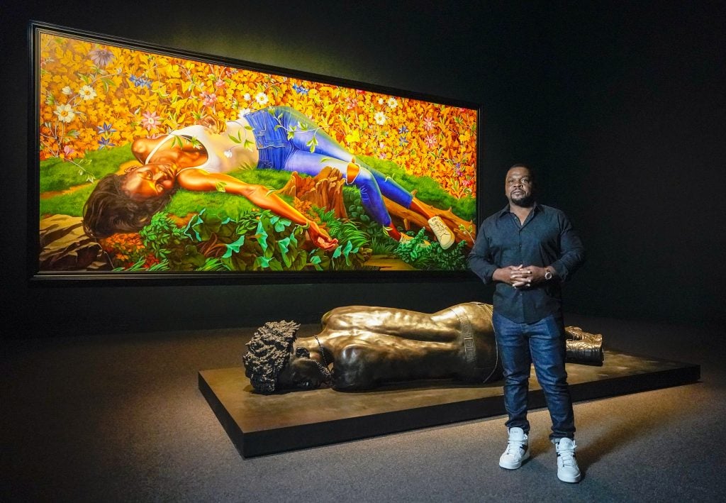 Artist Kehinde Wiley standing in front of one of his paintings and sculptures in a darkened gallery.
