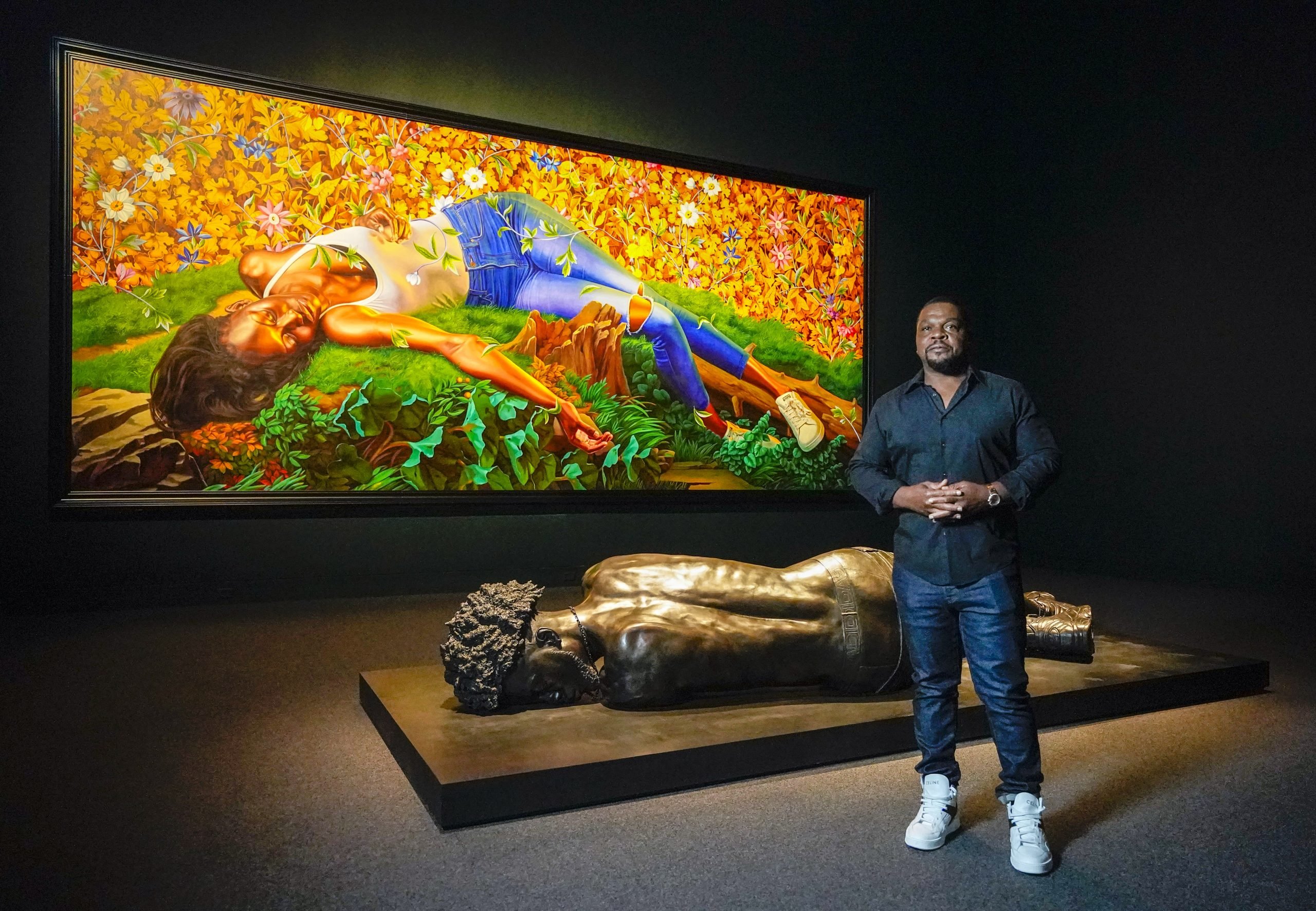 Artist Kehinde Wiley standing in front of one of his paintings and sculptures in a darkened gallery.