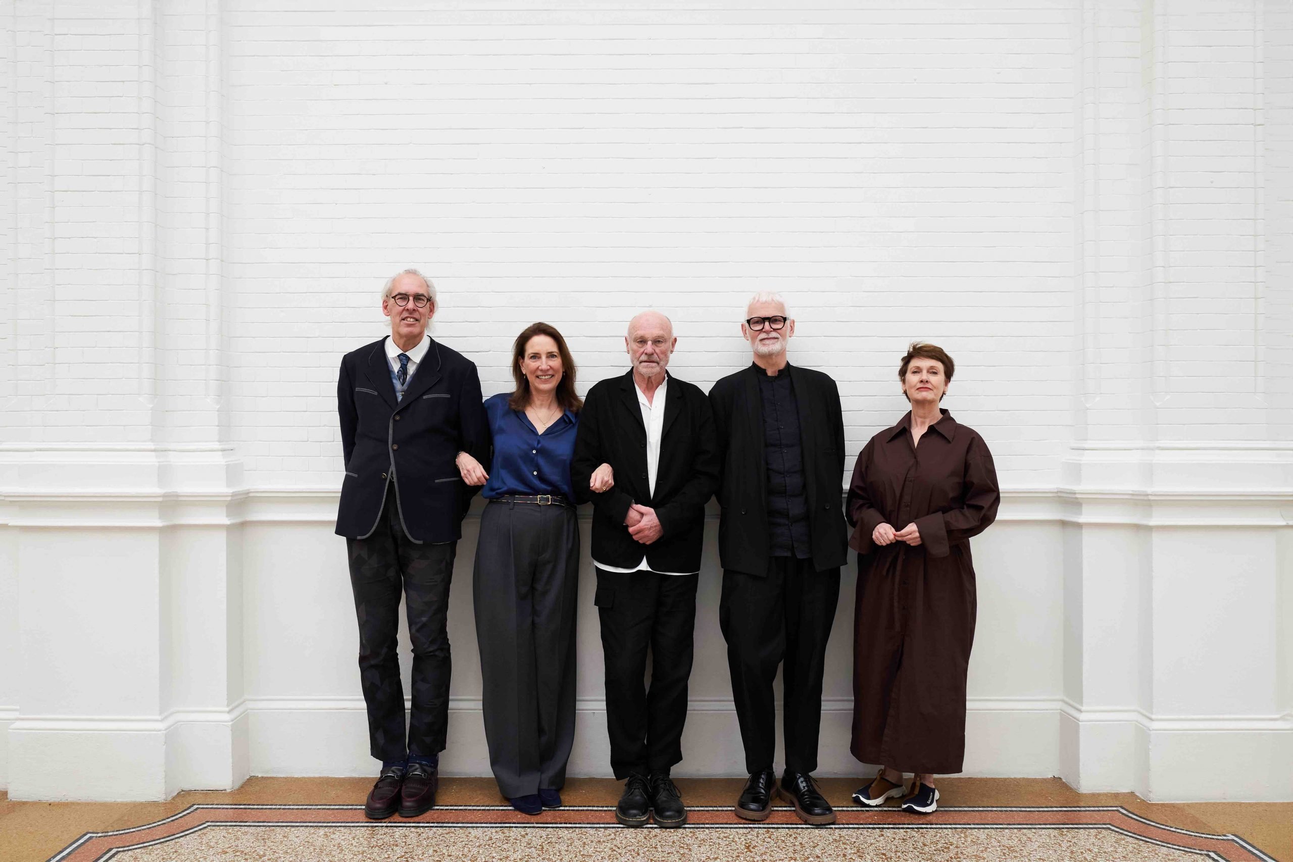 Five people, including three men and two women, standing against a white brick wall.