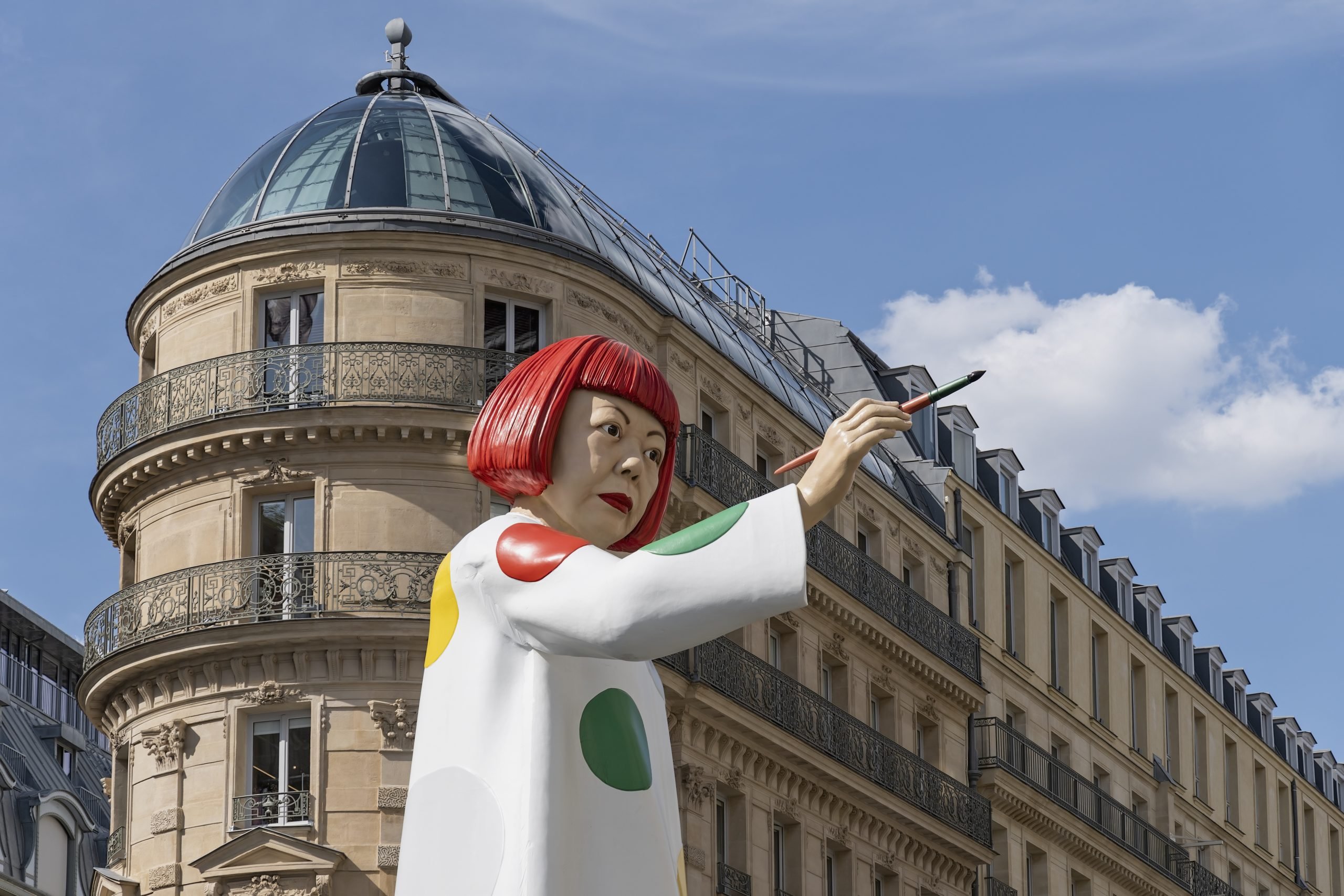 A huge sculpture in the likeness of Japanese artist Yayoi Kusama pointing at a Louis Vuitton store which is out of frame.