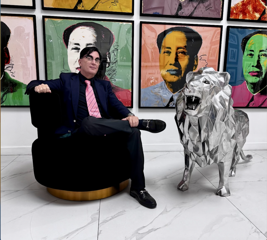 Portrait of Miami Fine Art Gallery founder Les Roberts sitting in front of a wall with multiple Andy Warhol prints of Mao Zedong sitting next to a silver chrome lion sculpture.
