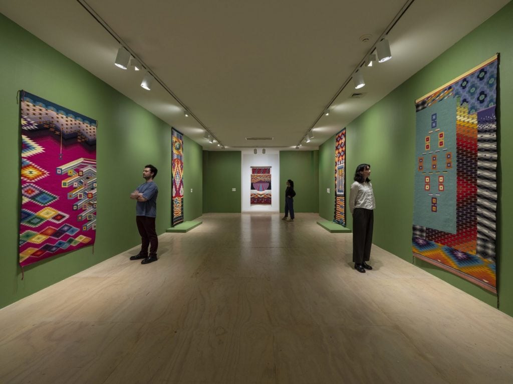 A gallery with green painted walls where people look at large complex textile hangings