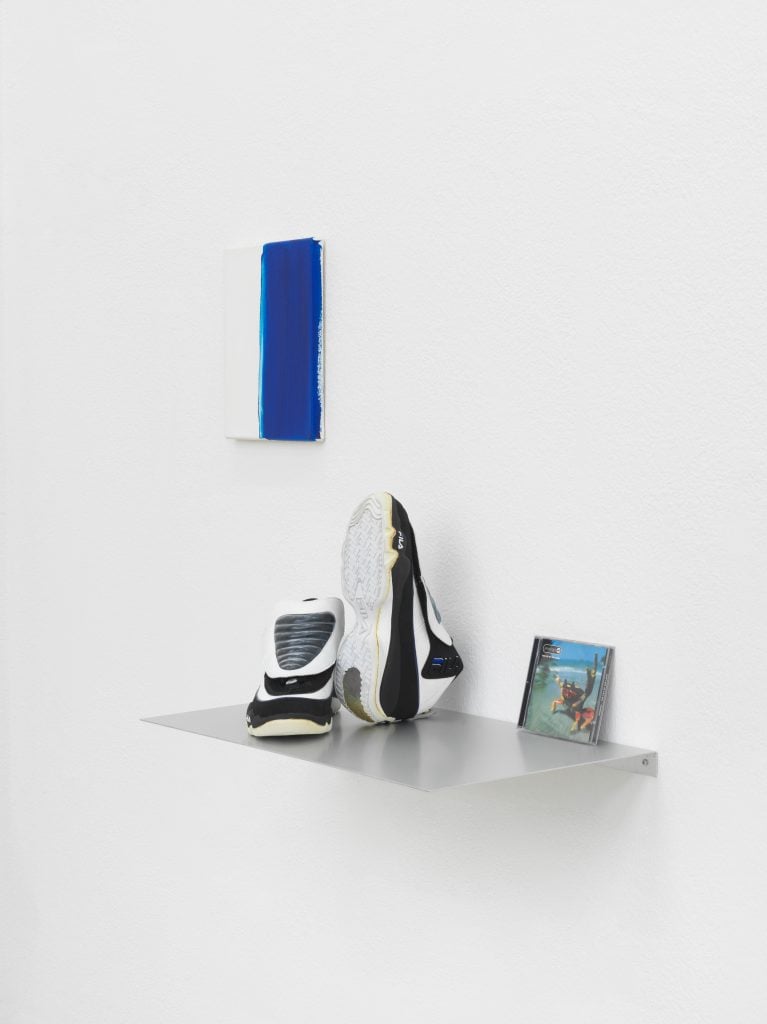 Sneakers, a small abstract blue and white painting and a CD are seen on a white wall