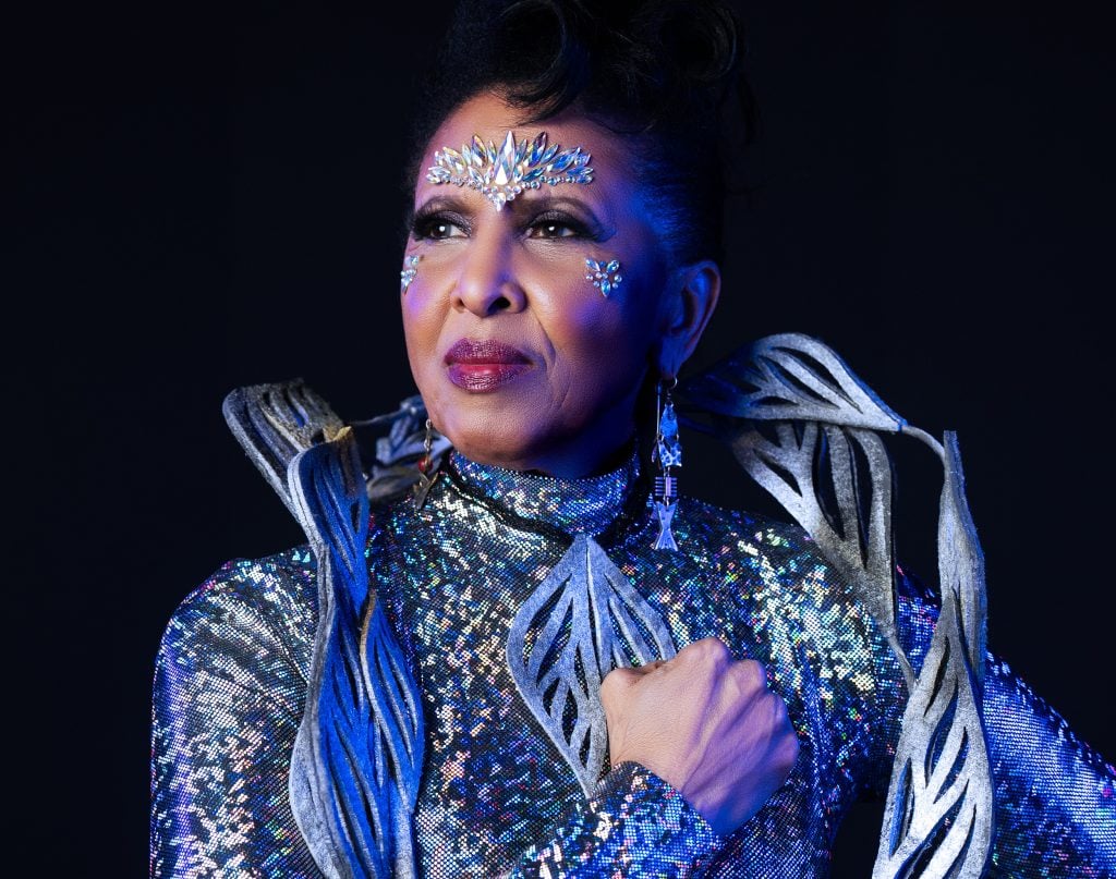 Artist Nona Hendryx dressed in a silver, futuristic jumpsuit, while holding her fist to her chest.