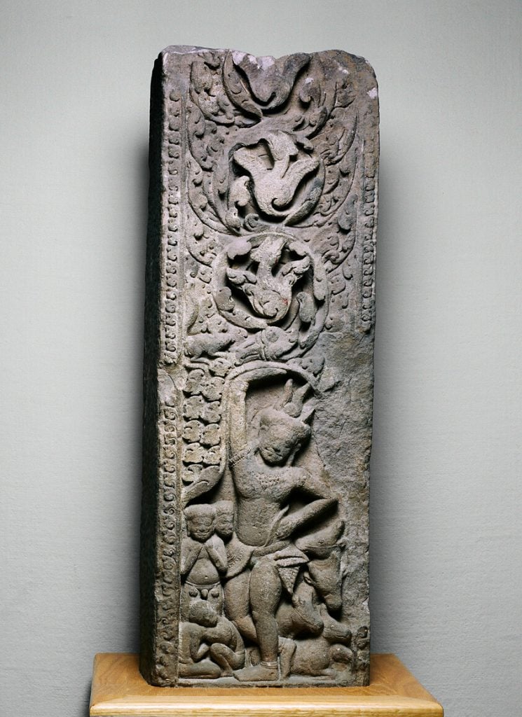 This image shows an ancient stone relief carving from the Phanom Rung historical site in Thailand. The relief is intricately detailed, depicting Hindu mythology scenes with figures in dynamic poses surrounded by decorative motifs and script. The carving is displayed on a plain background, highlighting its detailed artistry and craftsmanship.