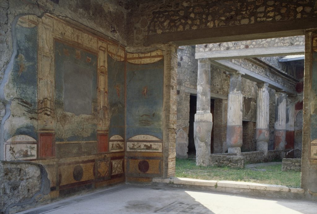 a large ancient house is covered in painted frescos and you can see outside there are ancient pillars surrounding a green grassy space