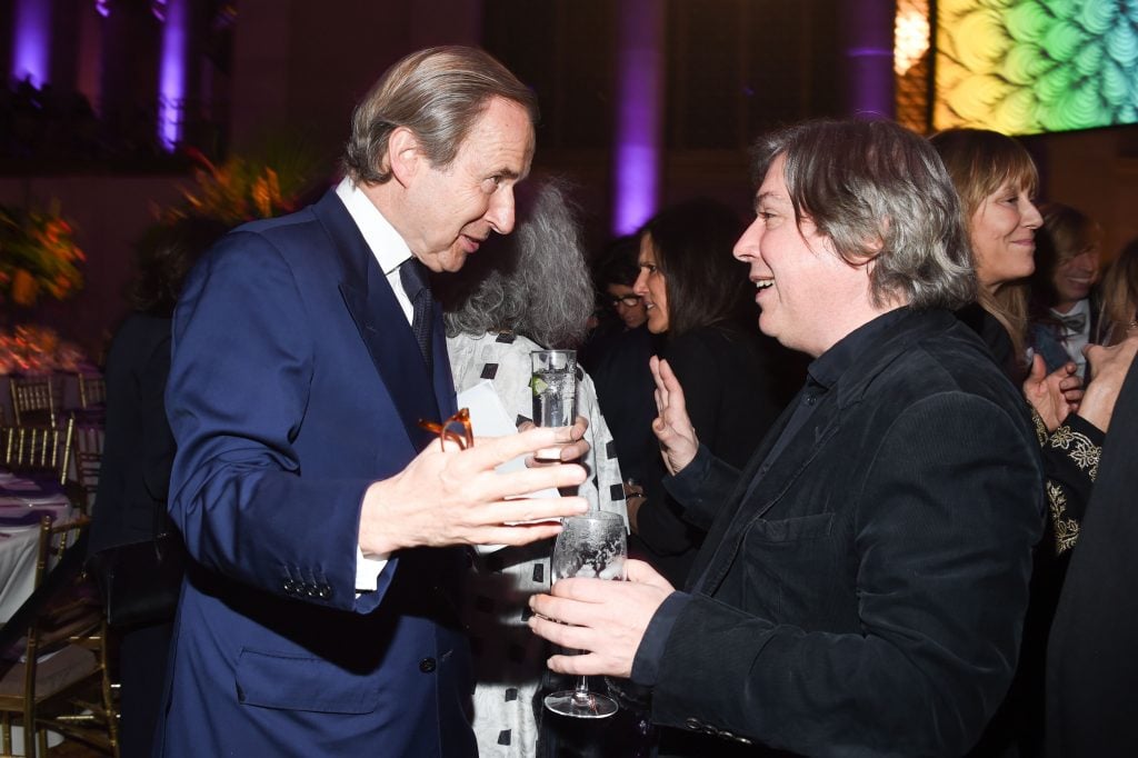 Two men, Simon de Pury and George Condo, speak animatedly to one another in a crowded room