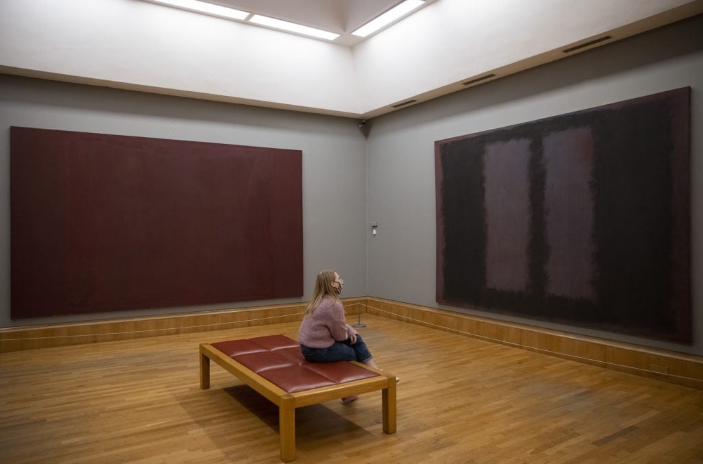 A figure sits on a bench in the middle of a gallery, gazing at a large abstract canvas