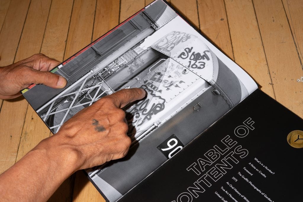 A person with a tattoo on their hand is pointing to graffiti art on a black-and-white photograph of a train car in an open book. The book's table of contents is visible on the right page, listing topics such as "The Graffiti Photographs of Gordon Matta-Clark" and "An Oral History of Writers." The background features a wooden floor.