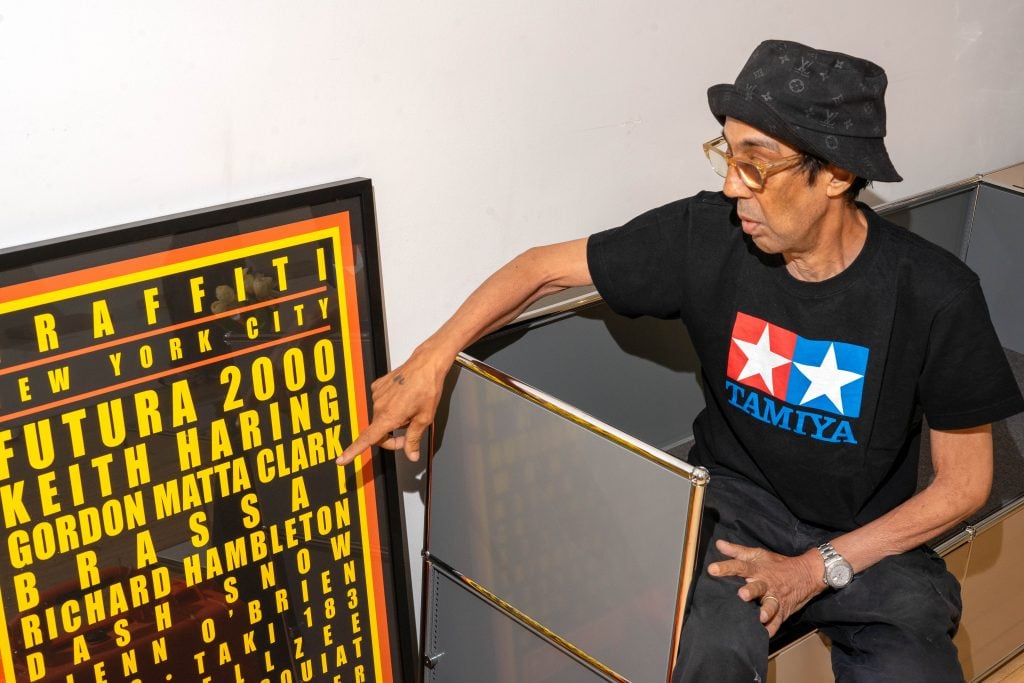 A man wearing a black Tamiya T-shirt and a black bucket hat is pointing at a framed poster listing names like Futura 2000, Keith Haring, and Gordon Matta Clark. He is sitting beside the poster, which appears to be related to a graffiti or street art exhibition in New York City.