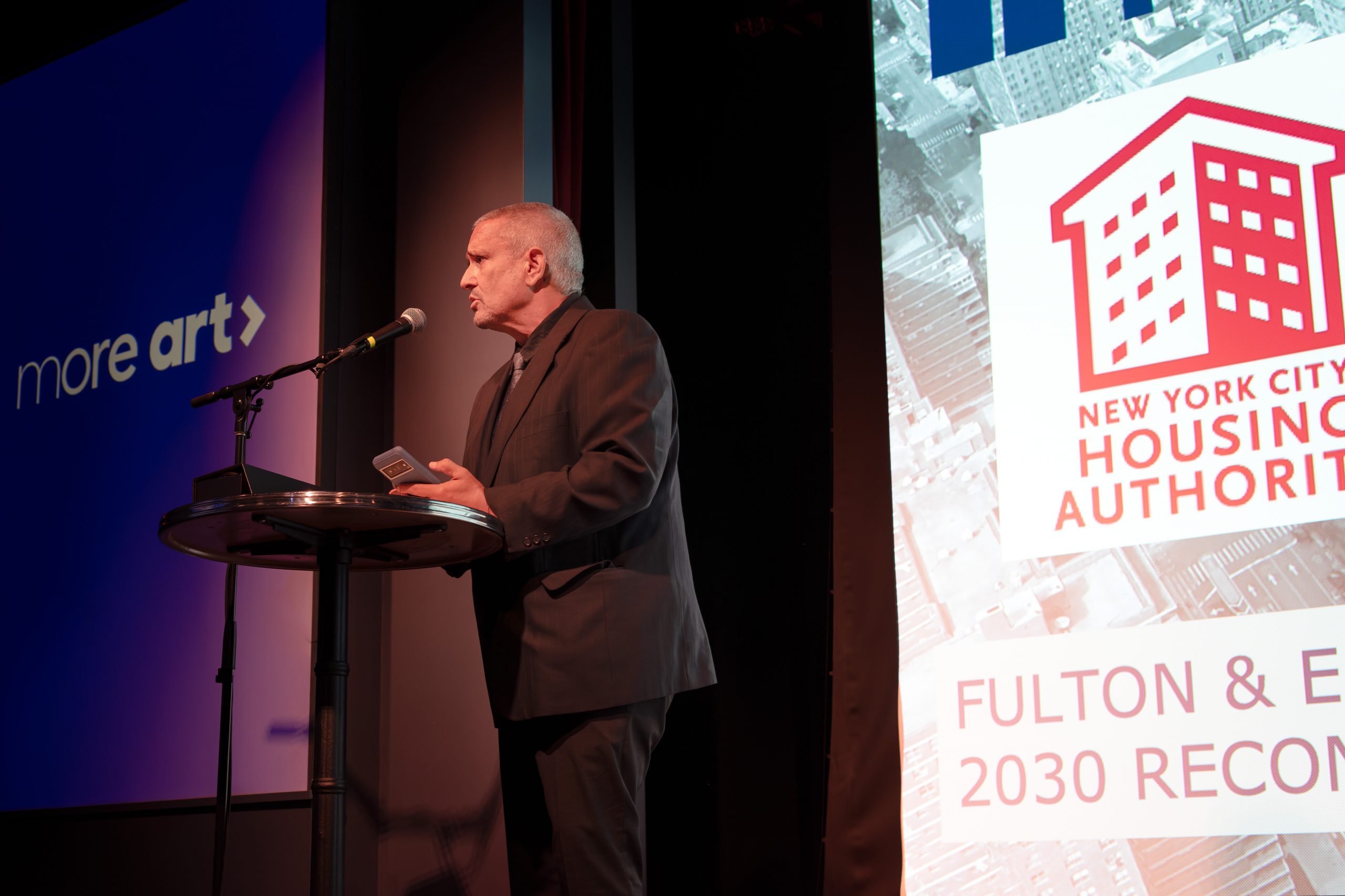 A man in a dark suit is speaking at a podium with a microphone. Behind him, there are two large screens. One screen displays the text "More Art," while the other screen shows the logo of the New York City Housing Authority and text about the Fulton and Elliott Chelsea Houses project. The man appears to be addressing an audience at an event.