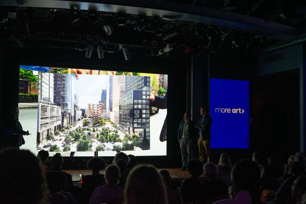 A large audience is seated in a darkened room, watching a presentation. The stage features a massive screen displaying a detailed architectural rendering of an urban development, with modern buildings and green spaces. Two men in suits stand to the right of the screen, observing the image. Another screen to the right displays the text "more art" on a blue background.
