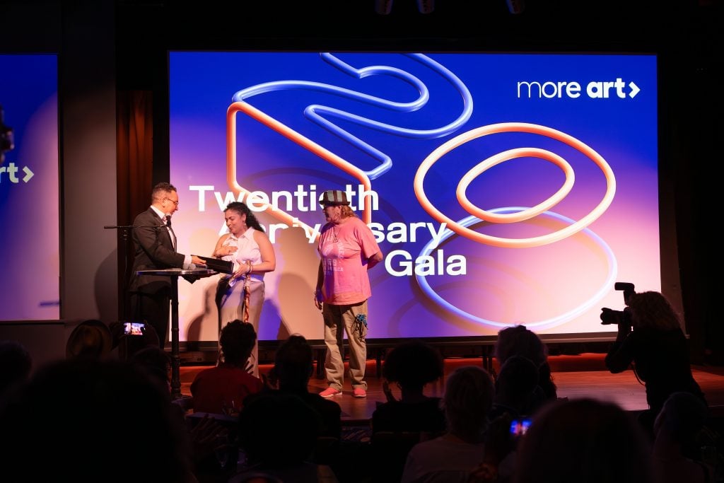 Three people are on stage at an event. One man in a suit is presenting an award or gift to a woman in a white dress, while another person in casual attire, including a pink t-shirt and hat, stands next to them. Behind them, a large screen displays the text "Twentieth Anniversary Gala" with the "More Art" logo. The audience is seated and watching the presentation, and a photographer is capturing the moment.