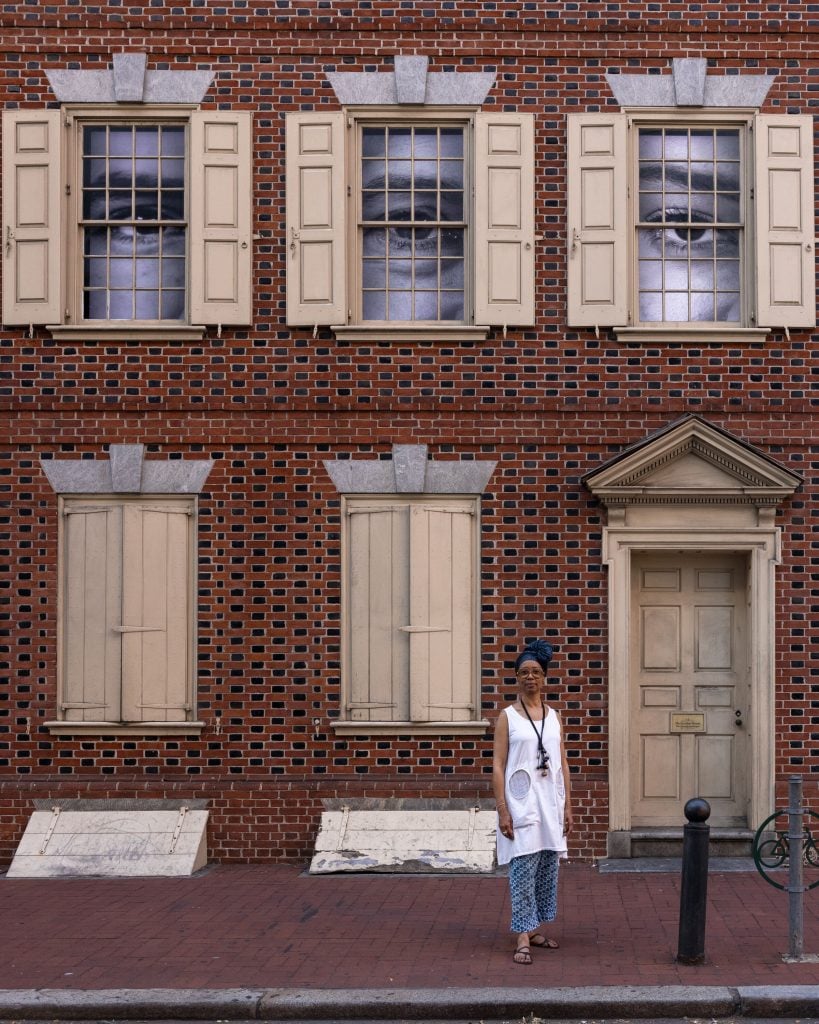 Artist Sonya Clark standing in front of a historic house with videos of eyes displayed out the windows