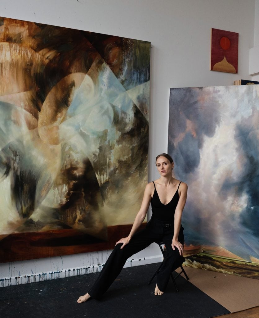 a white woman in a black leotard sits on a stool in front of two paintings of tumultous seascapes