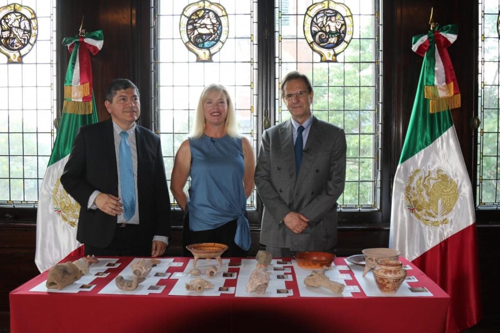 Two men and a woman standing in front of a table laden with Mayan artifacts