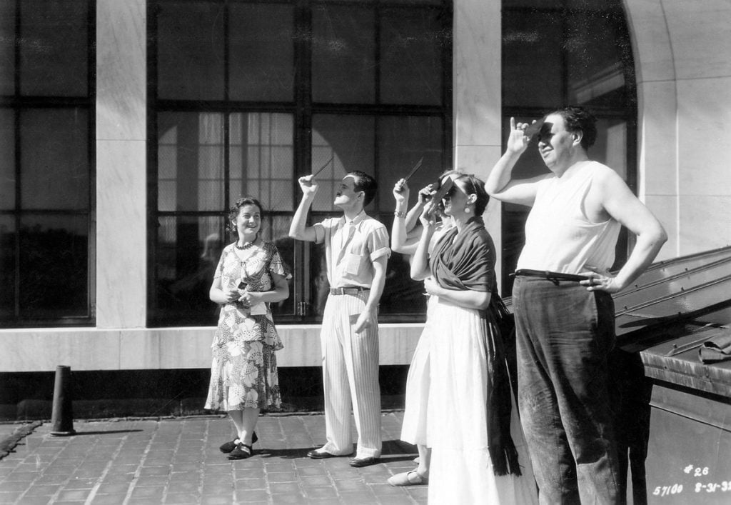 a black-and-white photograph of a group of men and women standing on a roof and looking into a solar eclipse