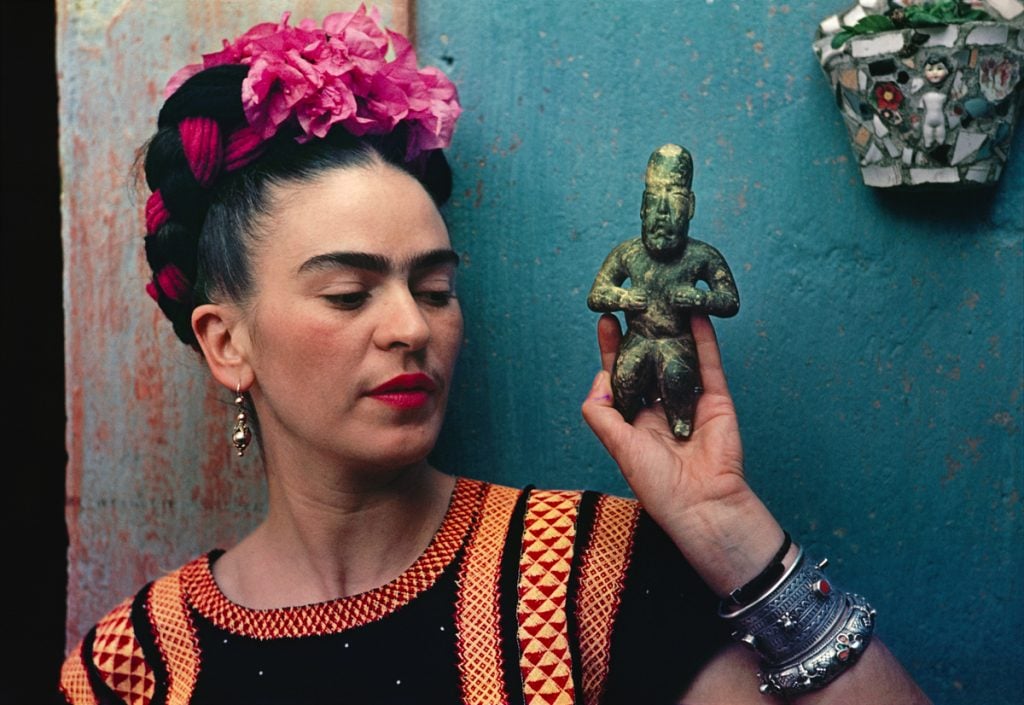 a color photograph of a woman with pink flower in her hair holding a small statuette in her hand in front of a turquoise wall