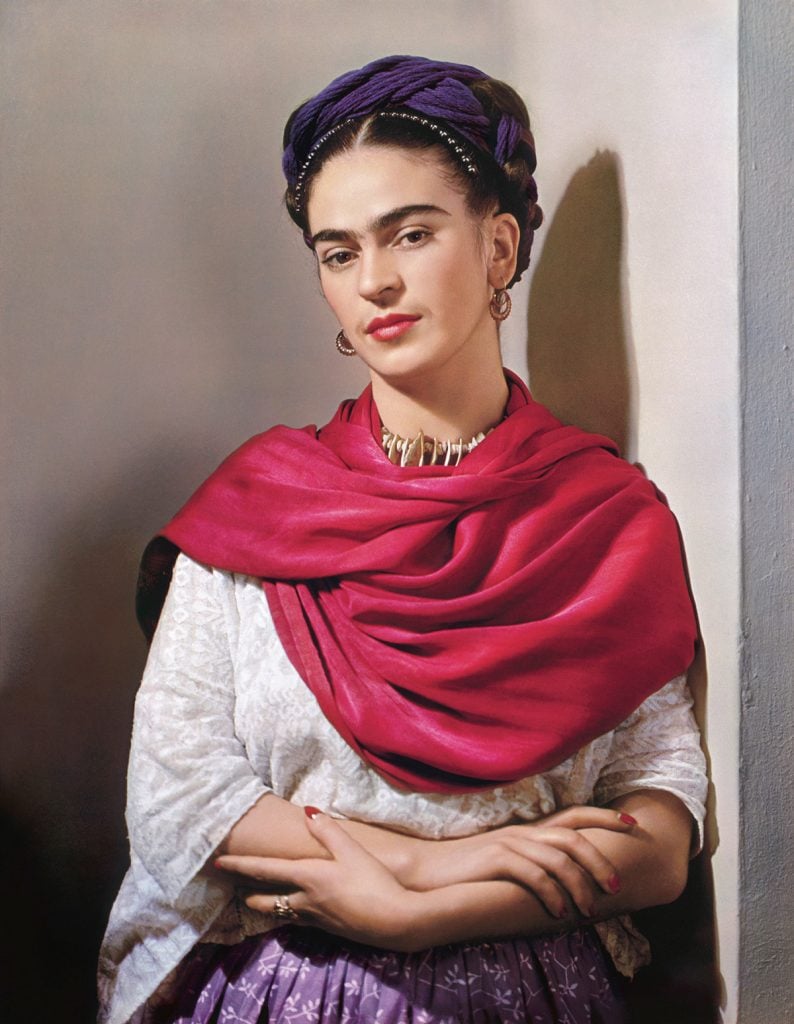 a woman wears purple flowers in her braided black hair, a fuschia pashmina, and aztec style jewelry, she poses in front of a wall with her hands clasped