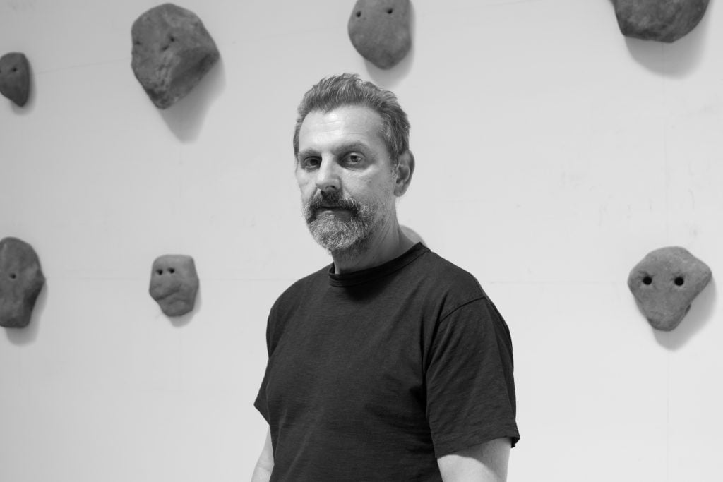 photograph of a man with a beard infront of small rock sculptures in the background
