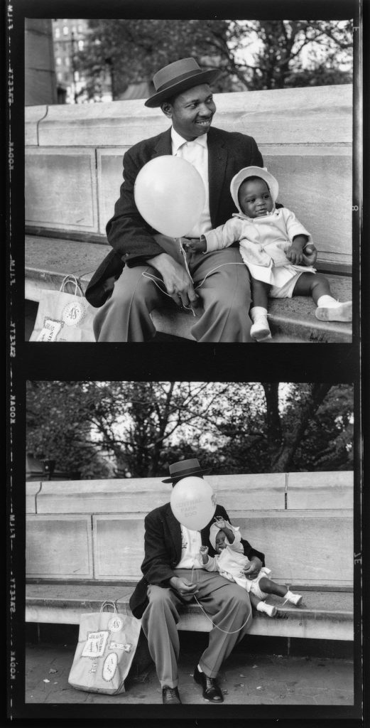 A Black man sits on a park bench with his small child who holds a balloon