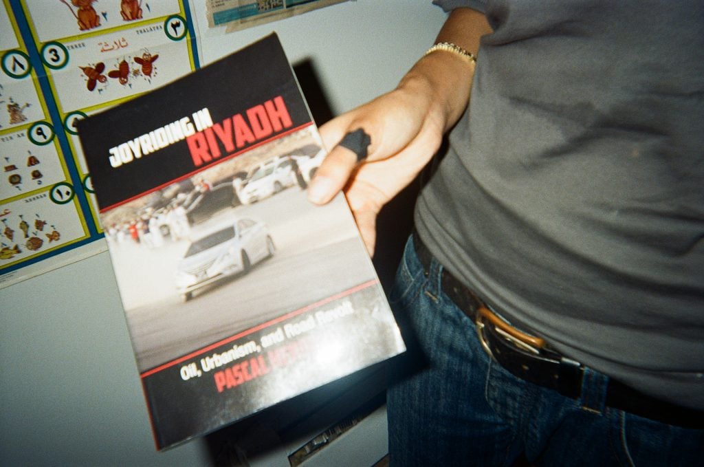 A woman's hand is seen holding a book titled 'Joyriding in Riyadh' that features an image of a car drifting on the cover.