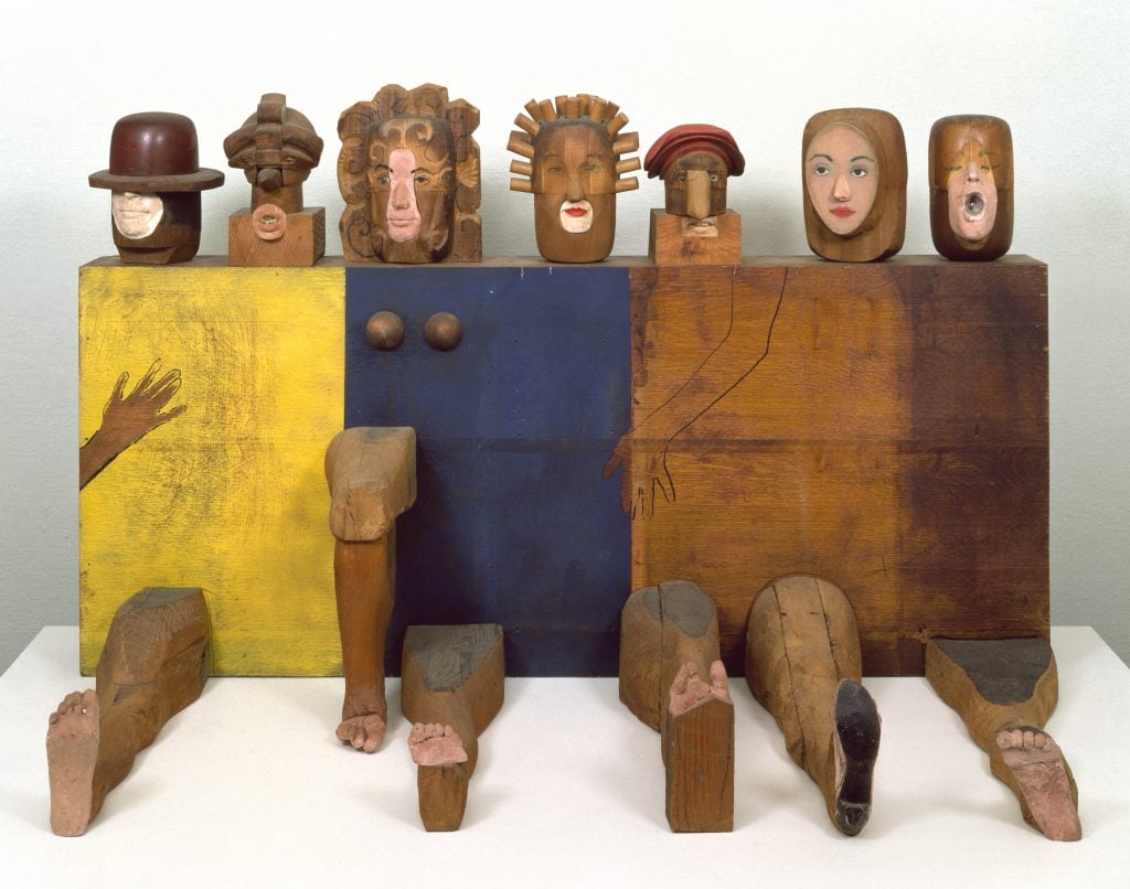 A blocky wooden Marisol sculpture of seven female heads on a rectangular body with six legs, one arm, and two breasts.