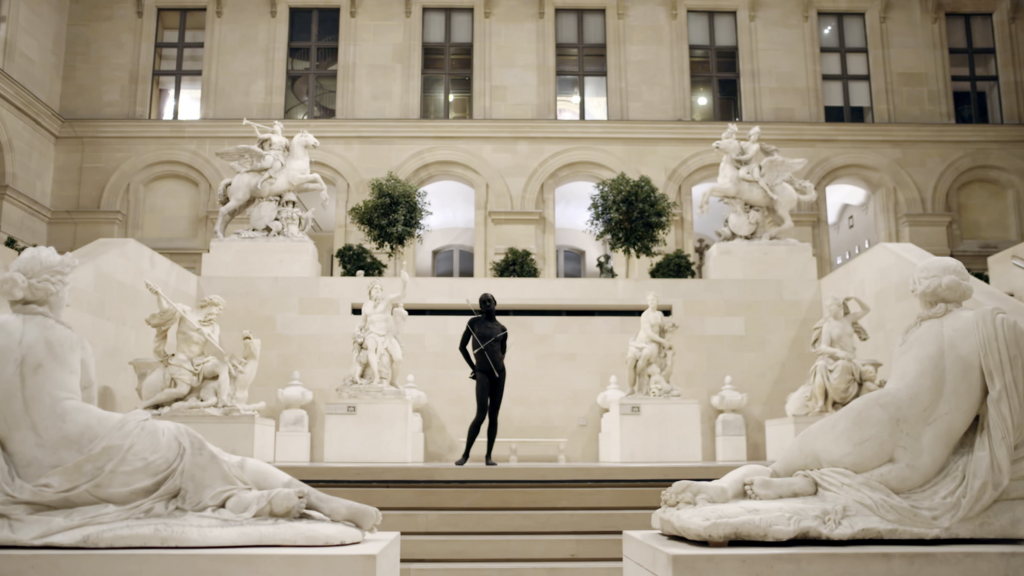 a black man stands in a contrapossto pose amidst white sculptures in similar positions