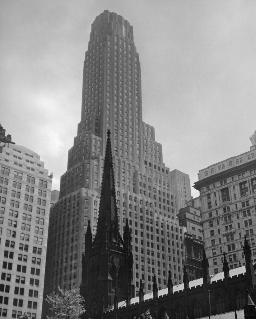 A black and white photograph of a 50-story Art Deco skyscraper in Lower Manhattan, with an ornate church steeple in the foreground. 