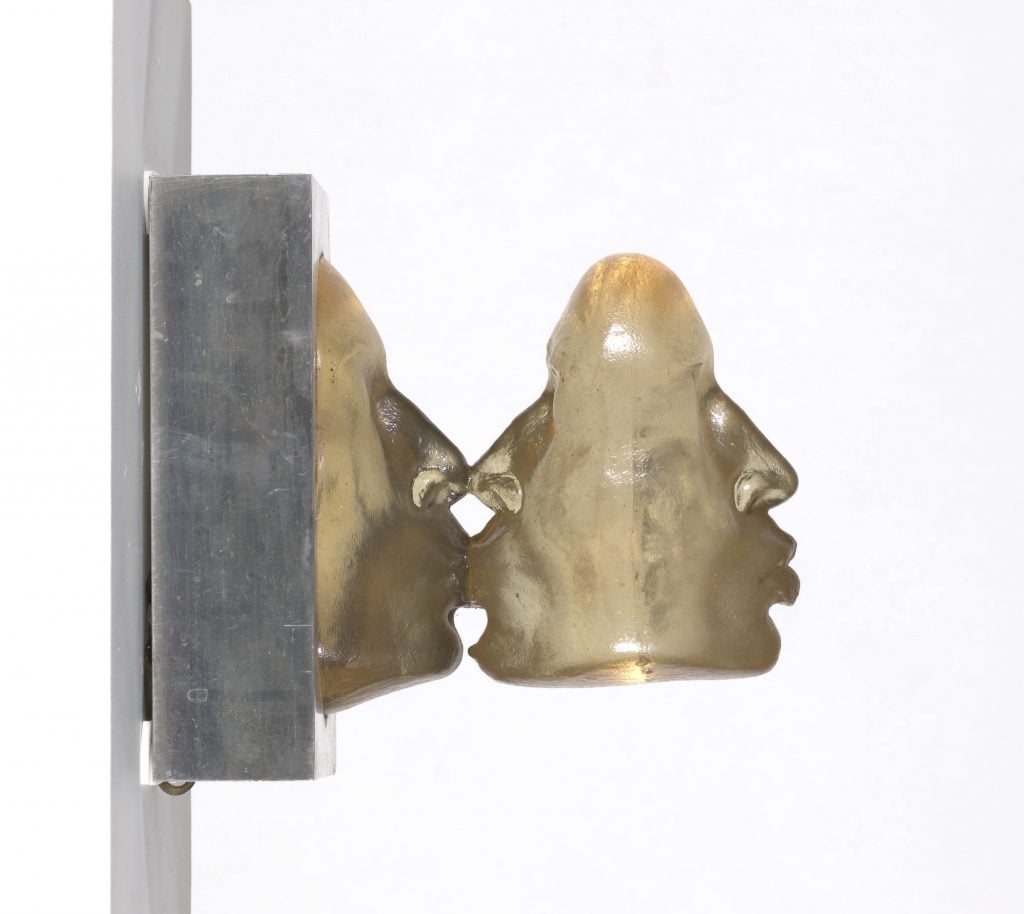 A surreal Marisol sculpture of three cast polyester faces, placed back to back on a metal base, mounted on the wall. Two of the faces are kissing, and the third grows out of the back of the second. 