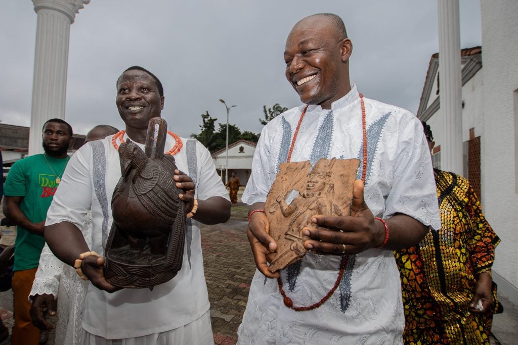 Two men in traditional white attire, adorned with coral bead necklaces, smile as they hold intricately carved wooden and bronze artifacts. They are standing outdoors, possibly at a cultural or ceremonial event, with several other people and structures visible in the background.