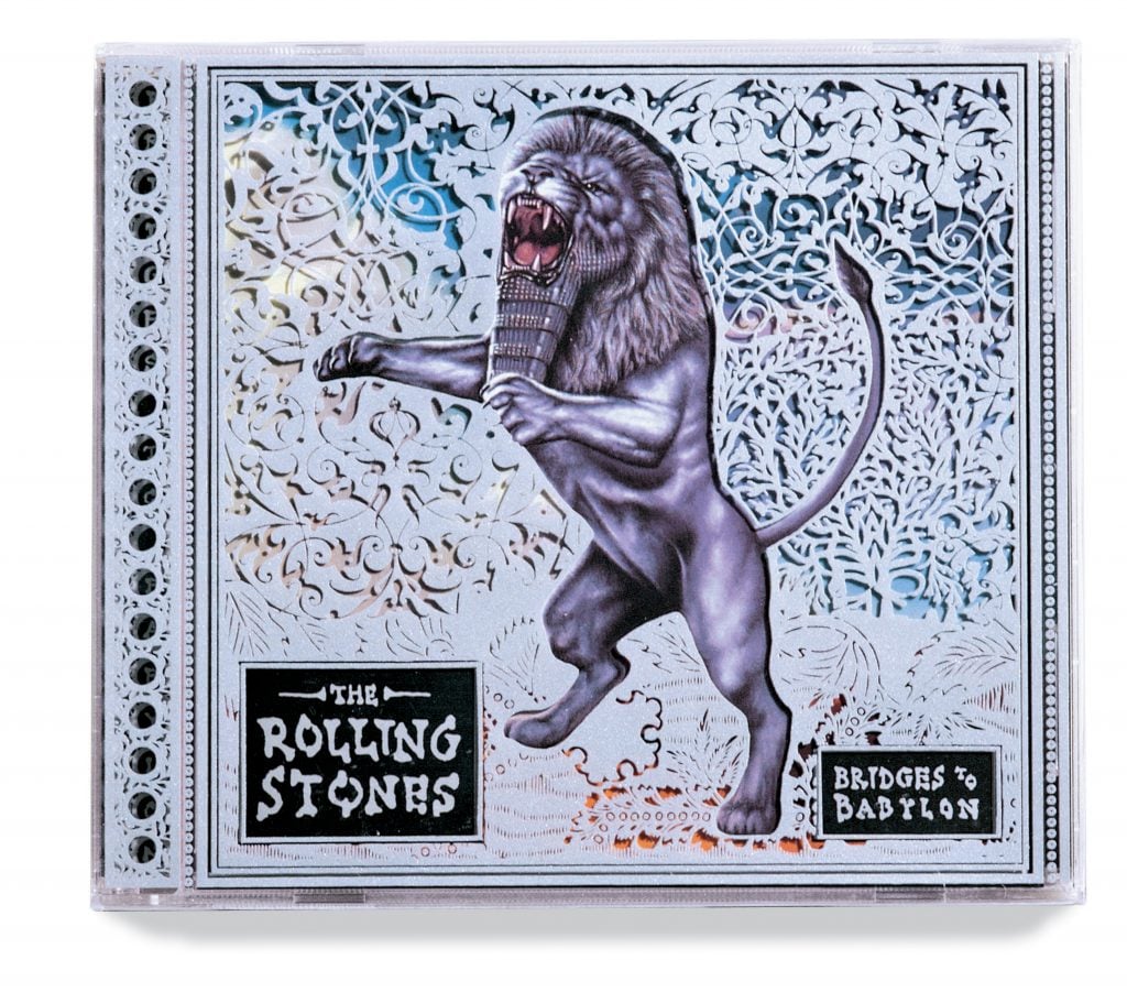 An album cover for "Bridges to Babylon" by The Rolling Stones, designed by Stefan Sagmeister. The cover features a stylized image of a roaring lion standing on its hind legs, holding a scepter. The background is intricately detailed with ornate patterns, giving a regal and mythical feel. The band's name and album title are displayed in bold, black text within decorative frames at the bottom left and right corners