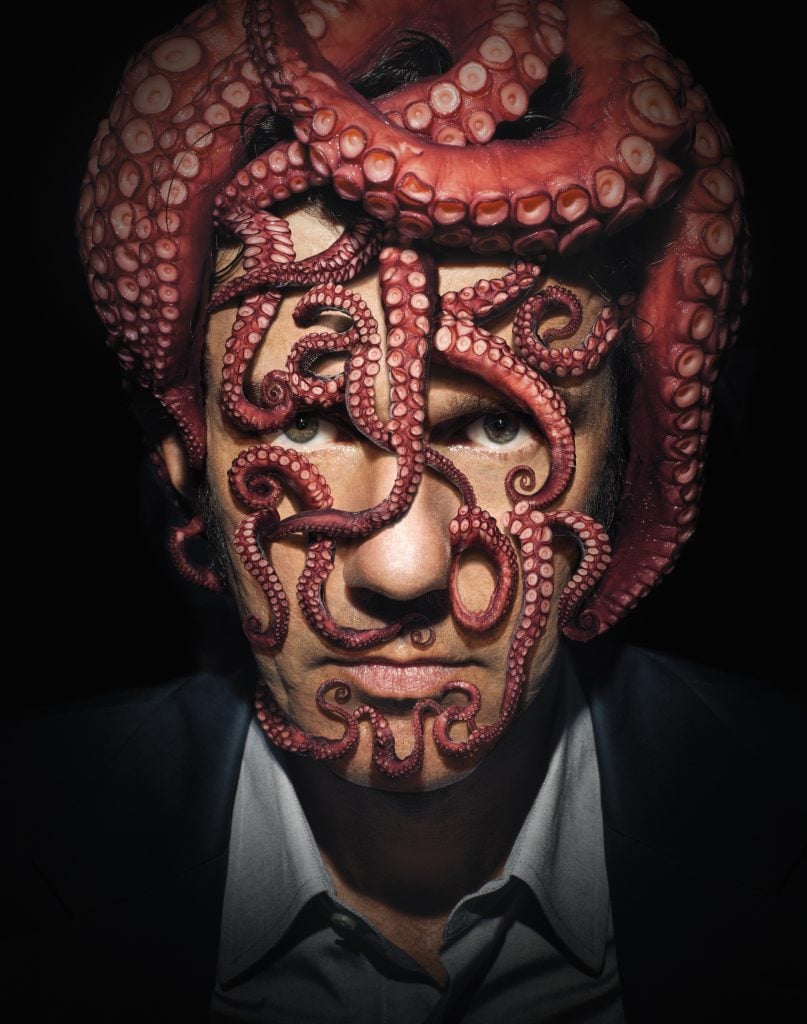 A print titled "Take It On" by Sagmeister & Walsh from 2013. The image features a close-up of a man's face, partially obscured by a large, coiled octopus whose tentacles wrap around his head and face. The tentacles are intricately arranged to form the words "Take It On." The man is dressed in a suit, and the dramatic lighting highlights the texture and detail of the octopus, creating a striking and surreal visual effect.