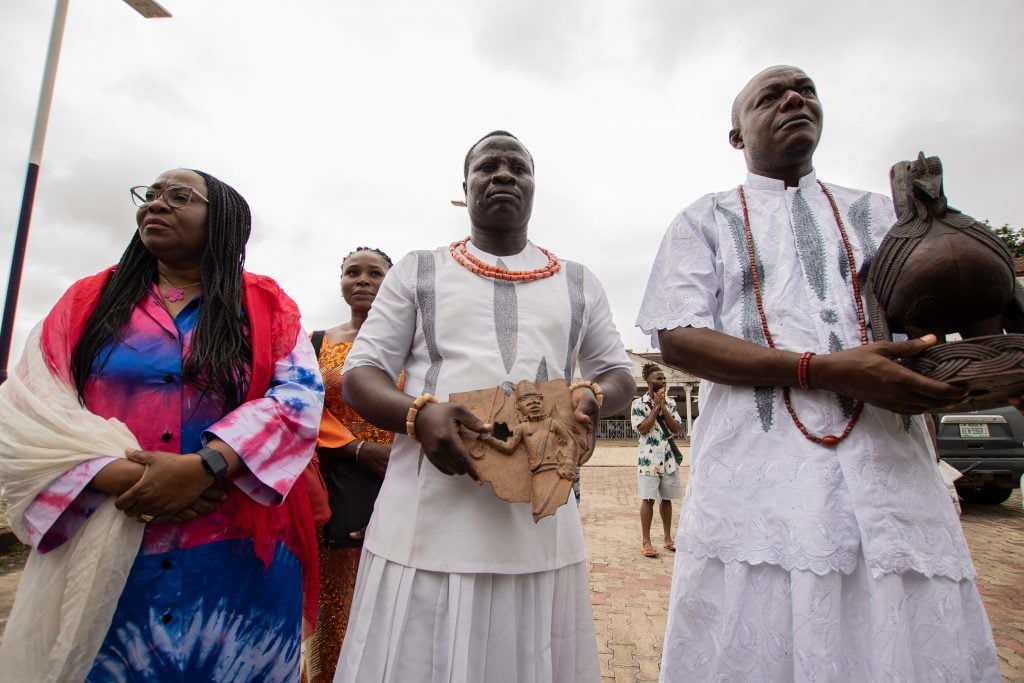 Three people stand solemnly outdoors: a woman in a colorful tie-dye outfit with a white shawl, and two men in traditional white attire adorned with coral beads, holding carved artifacts. Another woman in traditional attire stands behind them, and a few other people are seen in the background. The setting appears to be ceremonial.