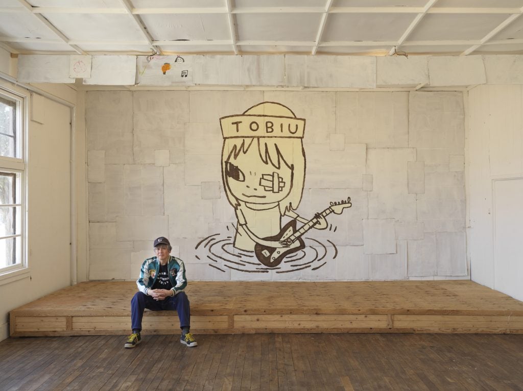 a man in casual clothes and a baseball hat sits in front of a large cartoonish drawing