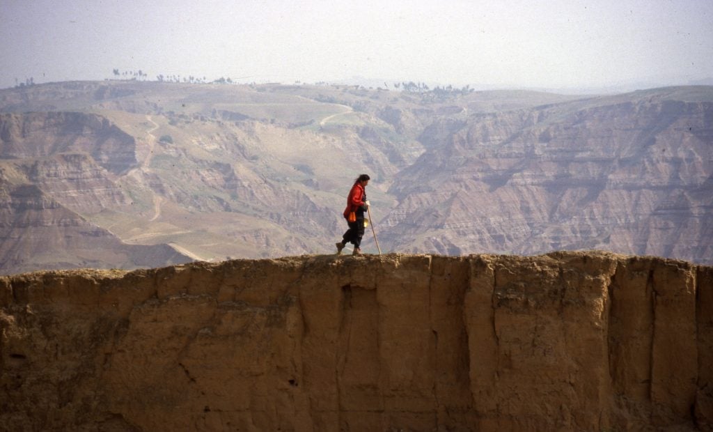 A young Marina Abramović is seen from a distance in a red jacket with a walking stick walking across a mountainous ridge during her 1988 performance piece with Ulay, "Great Wall Walk, China."