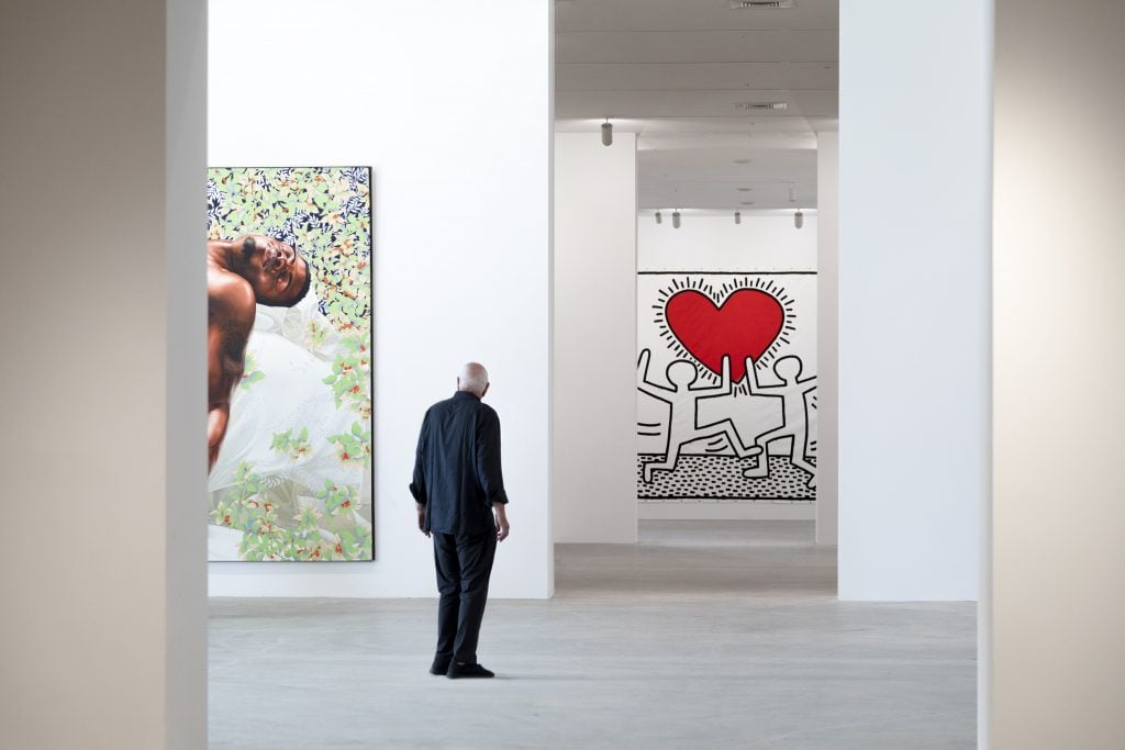 a man stands inside of a spacious museum with works by Keith Haring depicting a giant heart nearby 