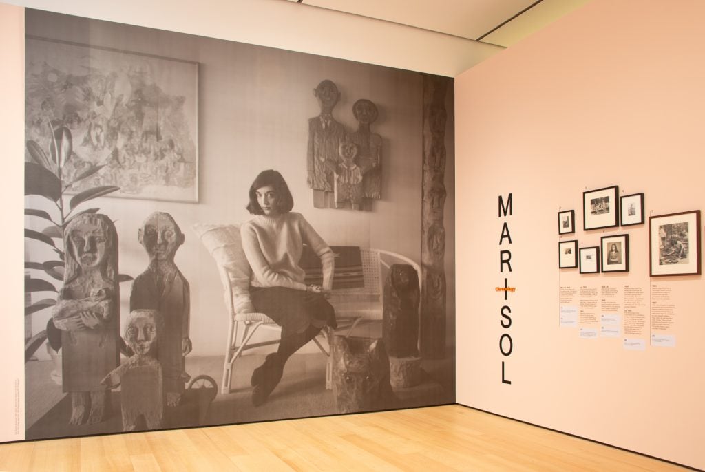 The entrance to "Marisol: A Retrospective," with one wall featuring a large black and white photo of the artist with her work as a young woman.
