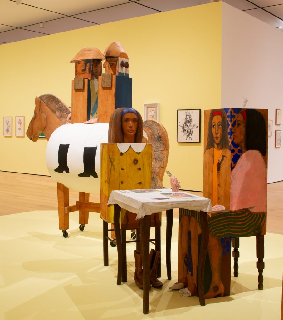 Wooden Marisol sculptures are on display in a museum gallery. A two figures each with the artist's face sit at a table, in front of a sculpture of two men in military uniforms riding on barrel horse.