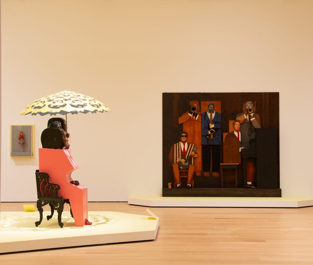 Wooden Marisol sculptures are on display in a museum gallery. A piece of two figures sitting with an umbrella on a bench rests on a platform, with a wooden relief sculpture of a jazz band with instruments is against the far wall. 