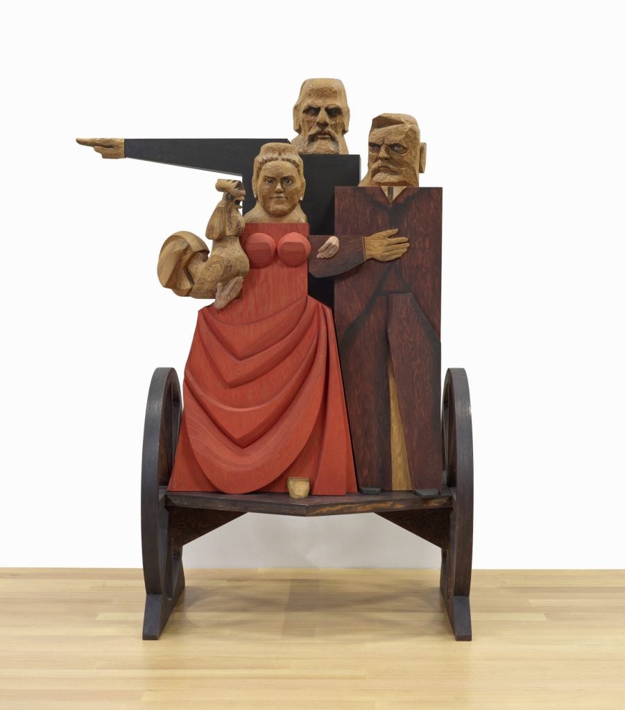 A large wooden Marisol sculpture of three people on a cart with two wheels. A woman in the left in a red dress holds a rooster, while the taller man behind her points to the side, and the man to her right holds a hand to his chest. The work is carved in a blocky, stylized fashion.