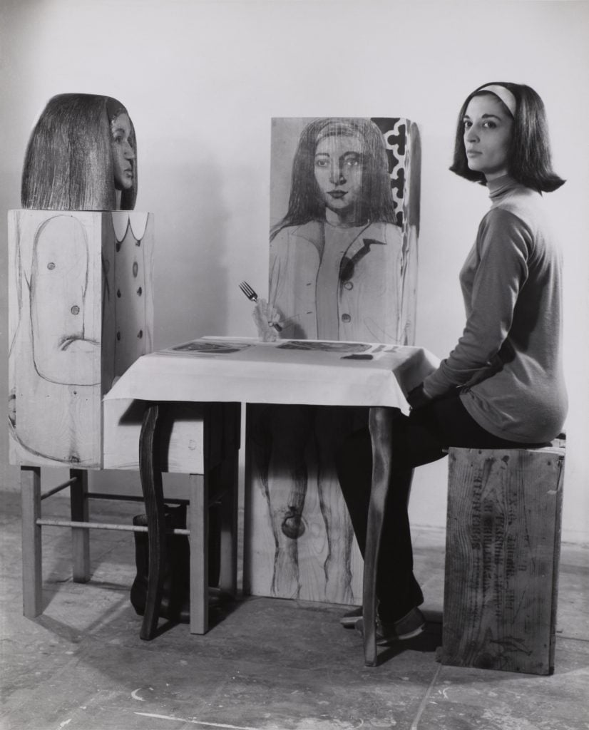 A black and white photograph of Marisol, a woman with a dark bob, sitting at her sculpture of a dinner table with two blocky wooden figures painted with her face. 