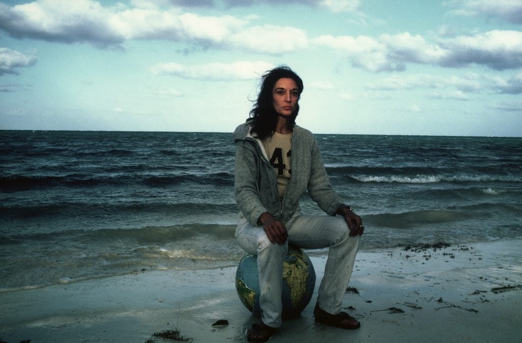 The artist Marisol sits on a globe on the shore of the ocean, dark blue waves behind her. She has dark, windswept hair that falls just below her shoulders, and wears light blue jeans and a gray zip up hoodie sweatshirt over an athletic t-shirt with the partially obscured number 43 on the front.