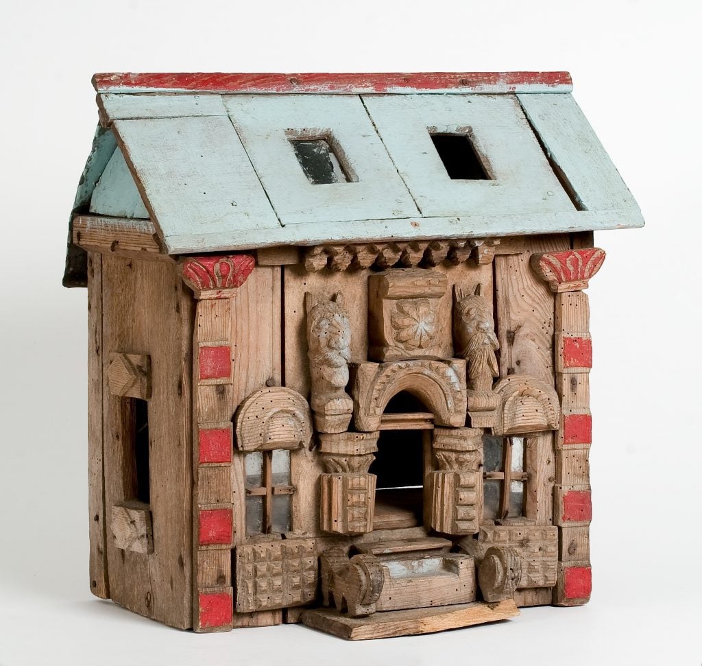A creation of art brut artist Auguste Forestier of a carved wooden house with incredible detail and carvings all across the surfaces and a pale green roof with two skylights.