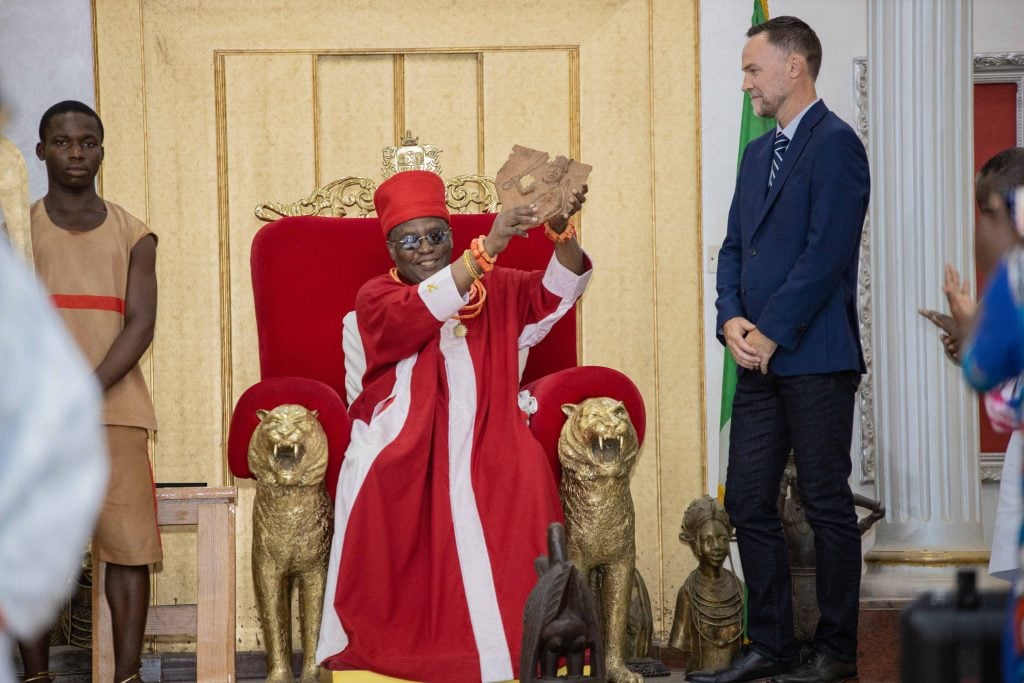 A regal figure, likely the Oba of Benin, dressed in red and adorned with coral beads, sits on a throne flanked by golden lion statues. He holds up an artifact while a young man in traditional attire stands beside him, holding a ceremonial staff. An observer in formal attire stands to the side. The setting is ornate, indicating a ceremonial event.