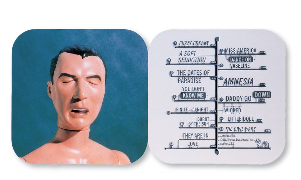 An image of the album booklet for David Byrne's "Feelings," designed by Stefan Sagmeister. The left side features a photo of a plastic mannequin head resembling Byrne with a pained expression, while the right side lists the album's track titles in a stylized format reminiscent of a mechanical display board. The design combines whimsical elements with a sleek, modern aesthetic.