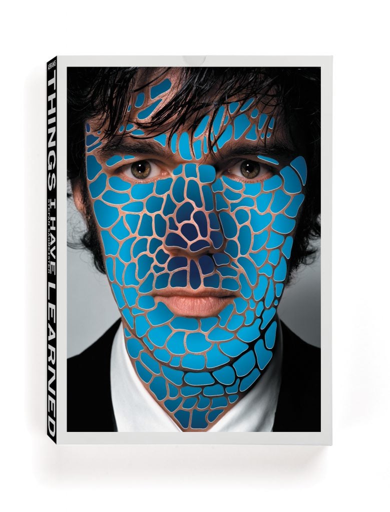 A book cover for Stefan Sagmeister's monograph "Things I Have Learned So Far In My Life." The cover features a close-up photograph of Sagmeister's face, partially obscured by a blue, mosaic-like pattern overlay. The spine of the book displays the title vertically in bold, black letters on a white background.