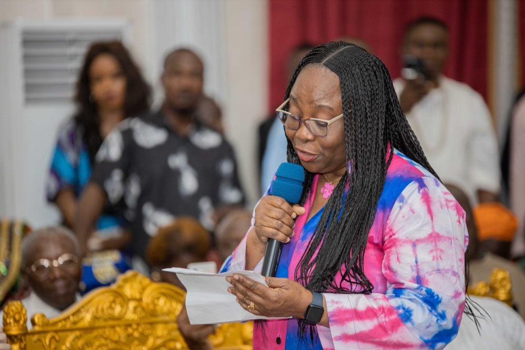 A woman in a colorful tie-dye outfit speaks into a microphone while reading from a paper at a formal event. She wears glasses and has long, braided hair. In the background, there are several people, some sitting and some standing, in what appears to be a ceremonial setting with ornate decorations.
