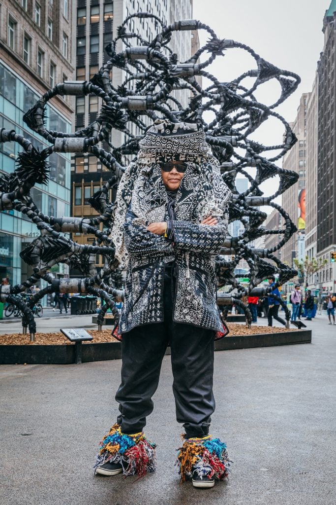 an older black woman wears an elaborate headdress. she stands on a city street in front of a towering black sculpture